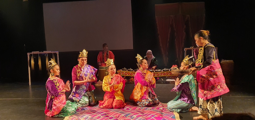 five ornately and brightly dressed phillipine people wearing shades of red green and gold with golden crowns on their heads, sit on a blanket hands in prayer. They seem to be attended by another figure, not crowned, carrying something to them in a basket. musicians on traditional instruments play in the background